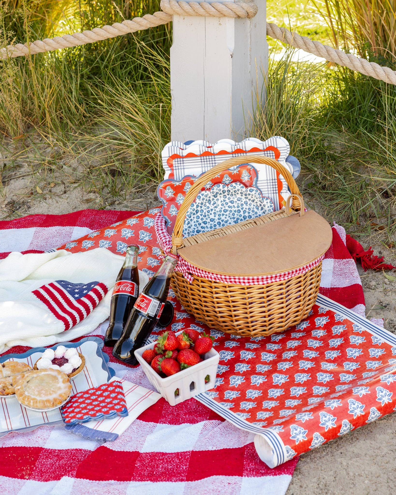 Beach picnic scene with red block print table runner and Americana paper plates in wicker basket on gingham blanket
