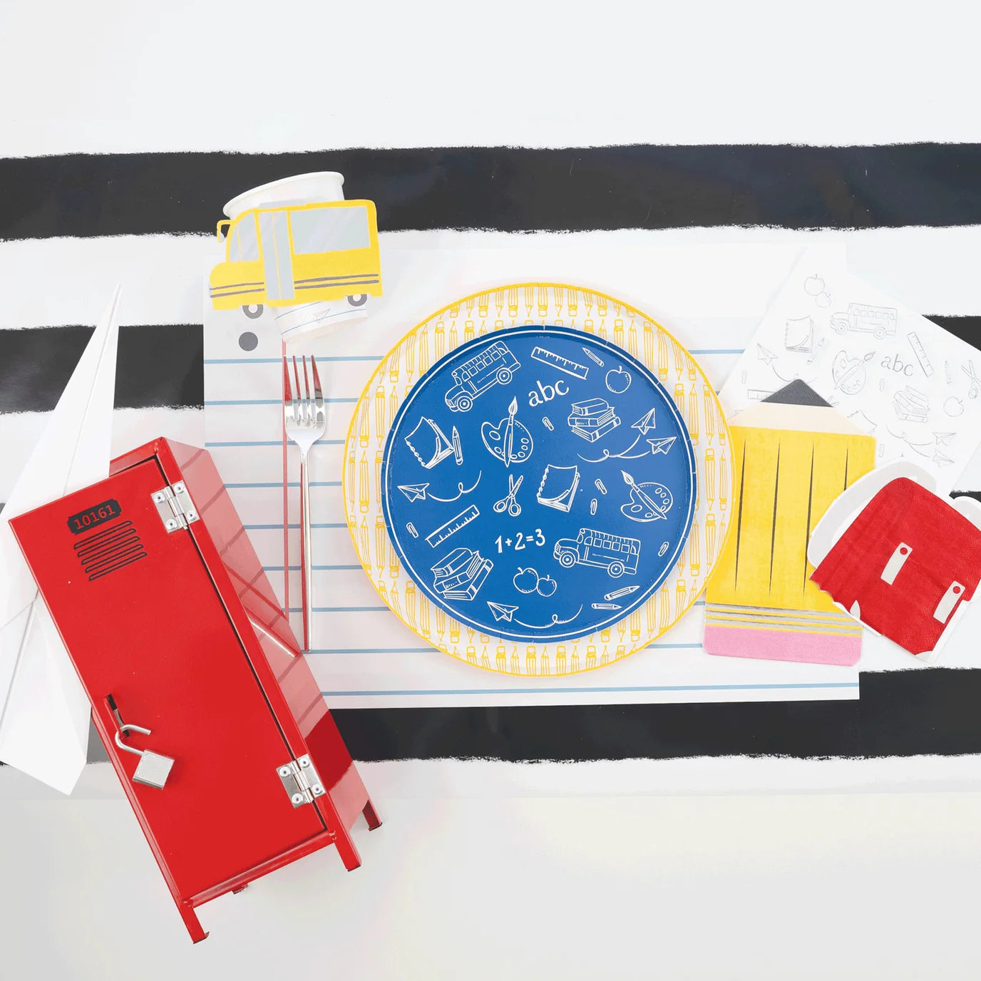Place setting with blue school‑icon plate nested in pencil dinner plate on notebook placemat, bus cup at top, red mini locker and pencil napkin alongside.