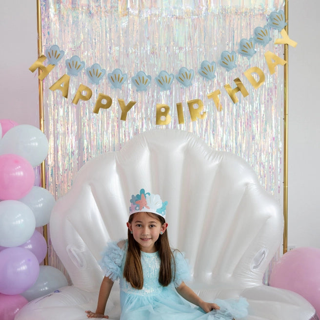 Full party scene: girl seated on giant white shell float, pastel balloons at side, banner duo overhead.
