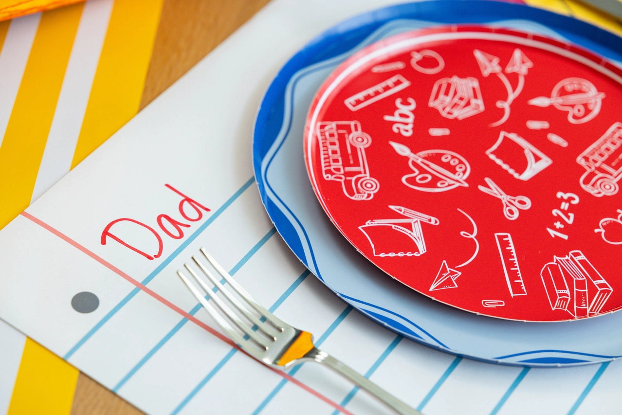 Close‑up of a place setting: red doodle plate over a blue rim plate, fork resting on a loose‑leaf placemat labeled “Dad” in red marker.