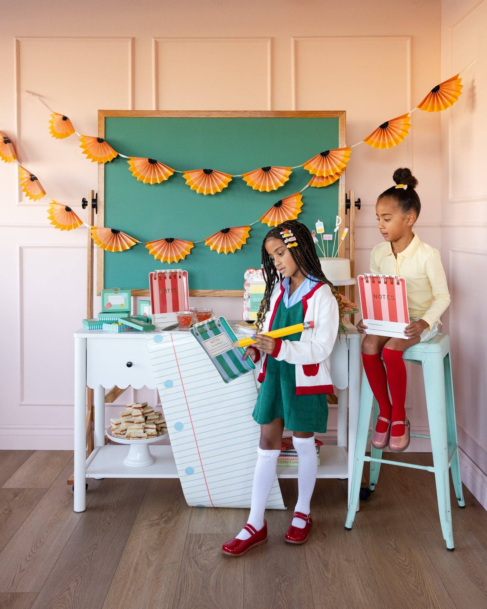 Two young girls at a styled back to school party with pencil shaving accordion banner, chalkboard backdrop, and notebook shaped paper plates displayed on a paper-scroll dessert cart