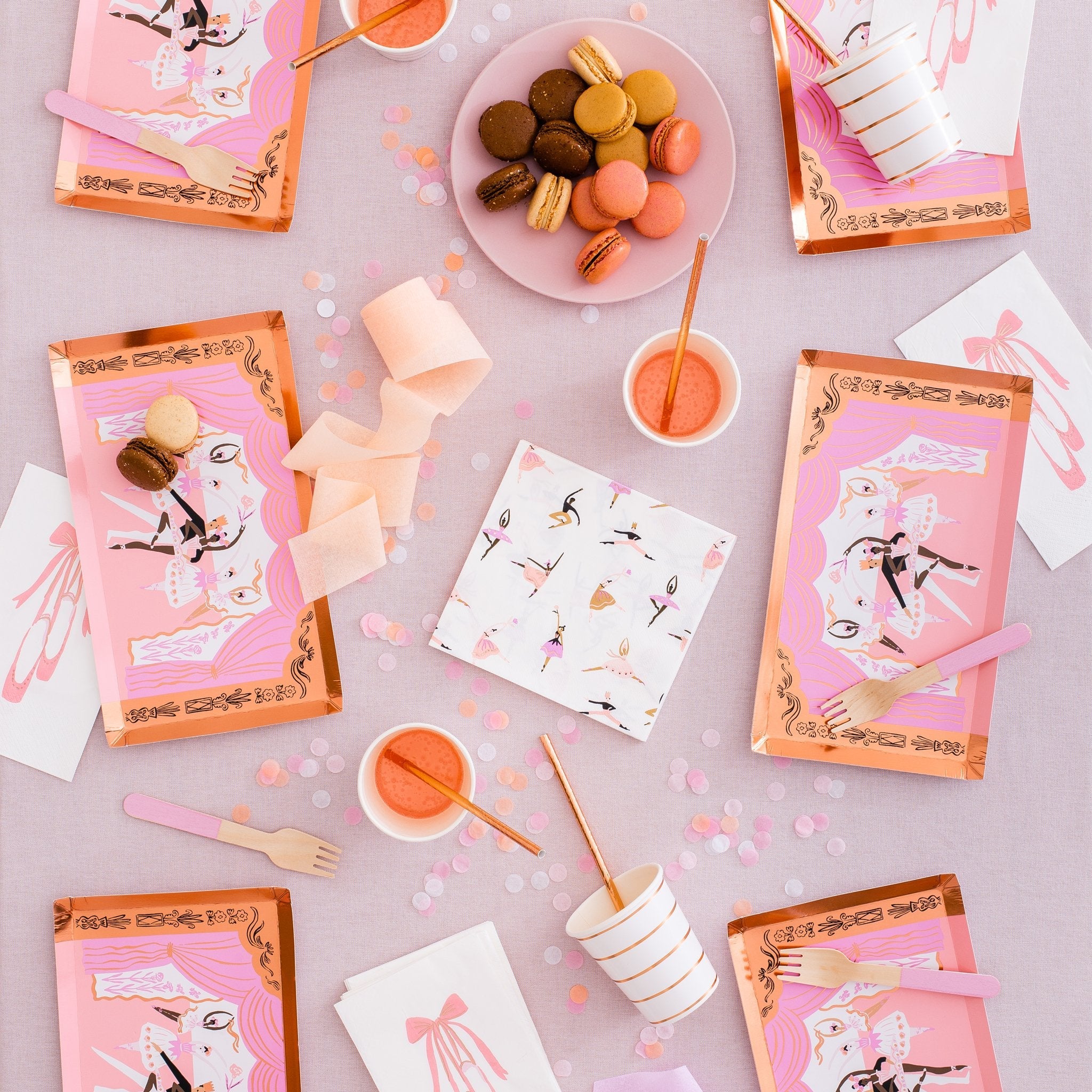 Overhead view of a pink ballet themed party table with stage-shaped plates, macarons, and gold striped cups.