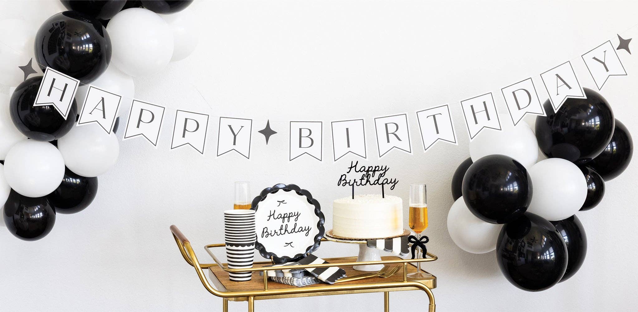 Black and white striped paper cups on gold bar cart with monochrome party supplies, black and white balloons, birthday cake, and banner