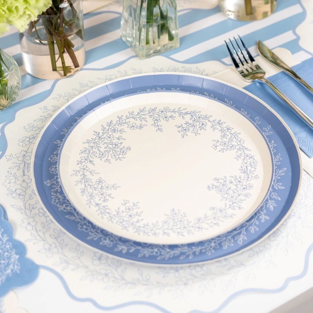 Dusty blue dinner plate with floral wreath design beside blue napkin and flatware.
