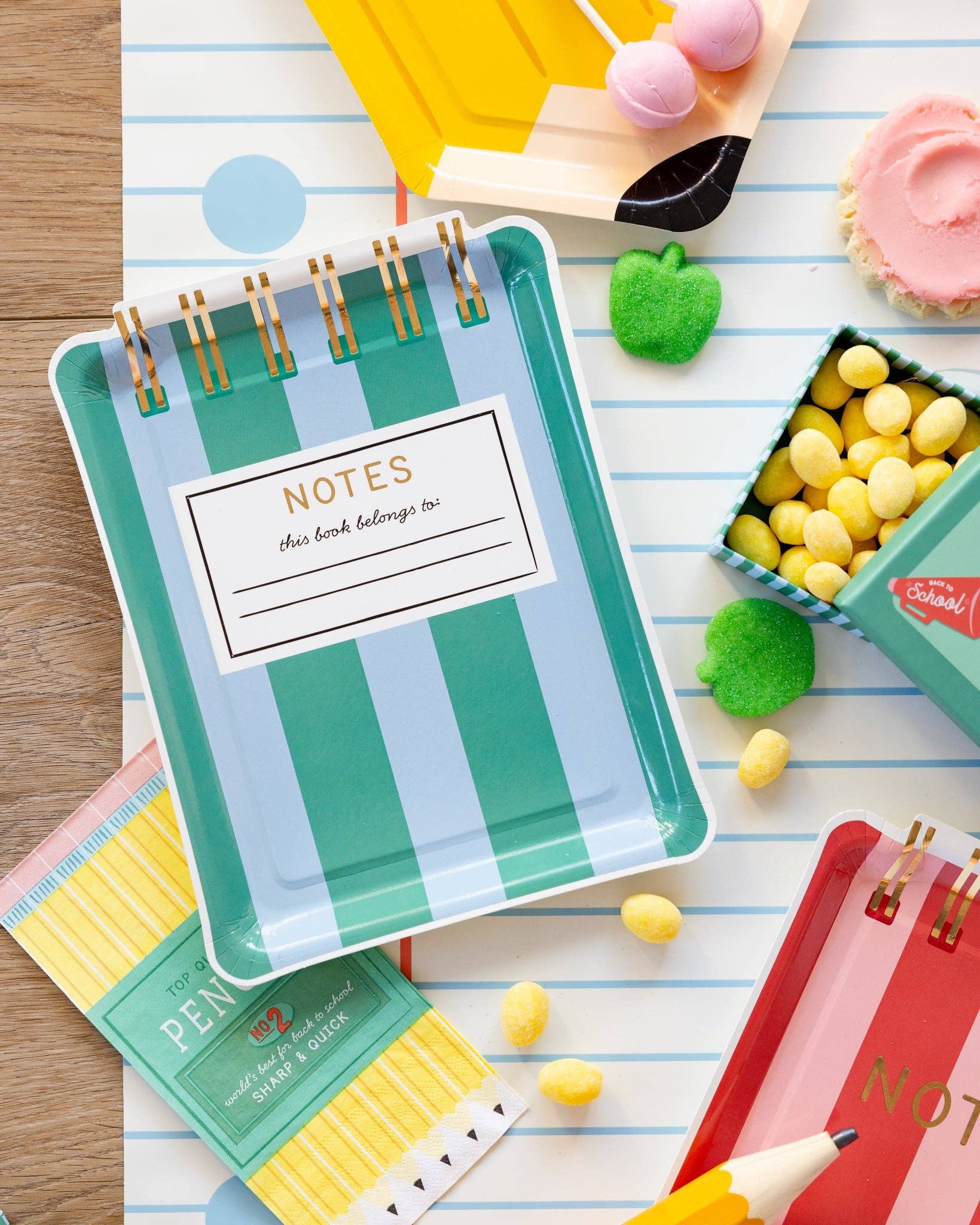 Flat lay of teal and blue striped notebook shaped paper plate on notebook paper runner with yellow candy, green apple candies, pencil shaped plate, and Back to School lunchbox treat box