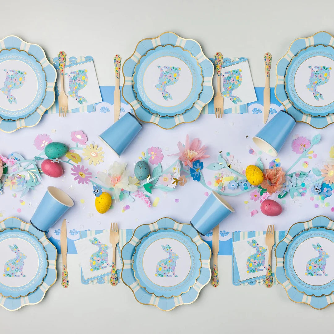 Blue striped scalloped paper guest napkins layered beneath floral bunny napkins on an Easter tablescape with blue scalloped paper plates, solid blue paper cups, floral wooden flatware, and a paper flower garland with speckled eggs.