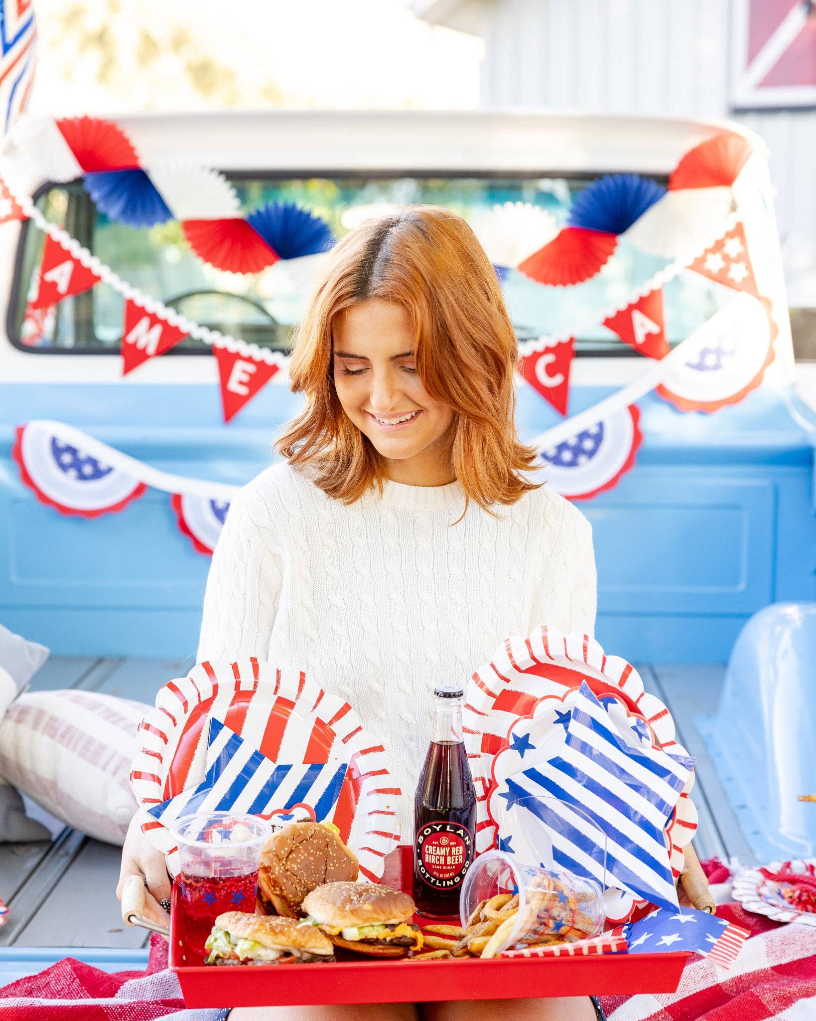 Lifestyle photo of woman holding a red tray loaded with burgers, blue striped star plates, red striped scalloped plates, and bottled sodas at an outdoor patriotic party
