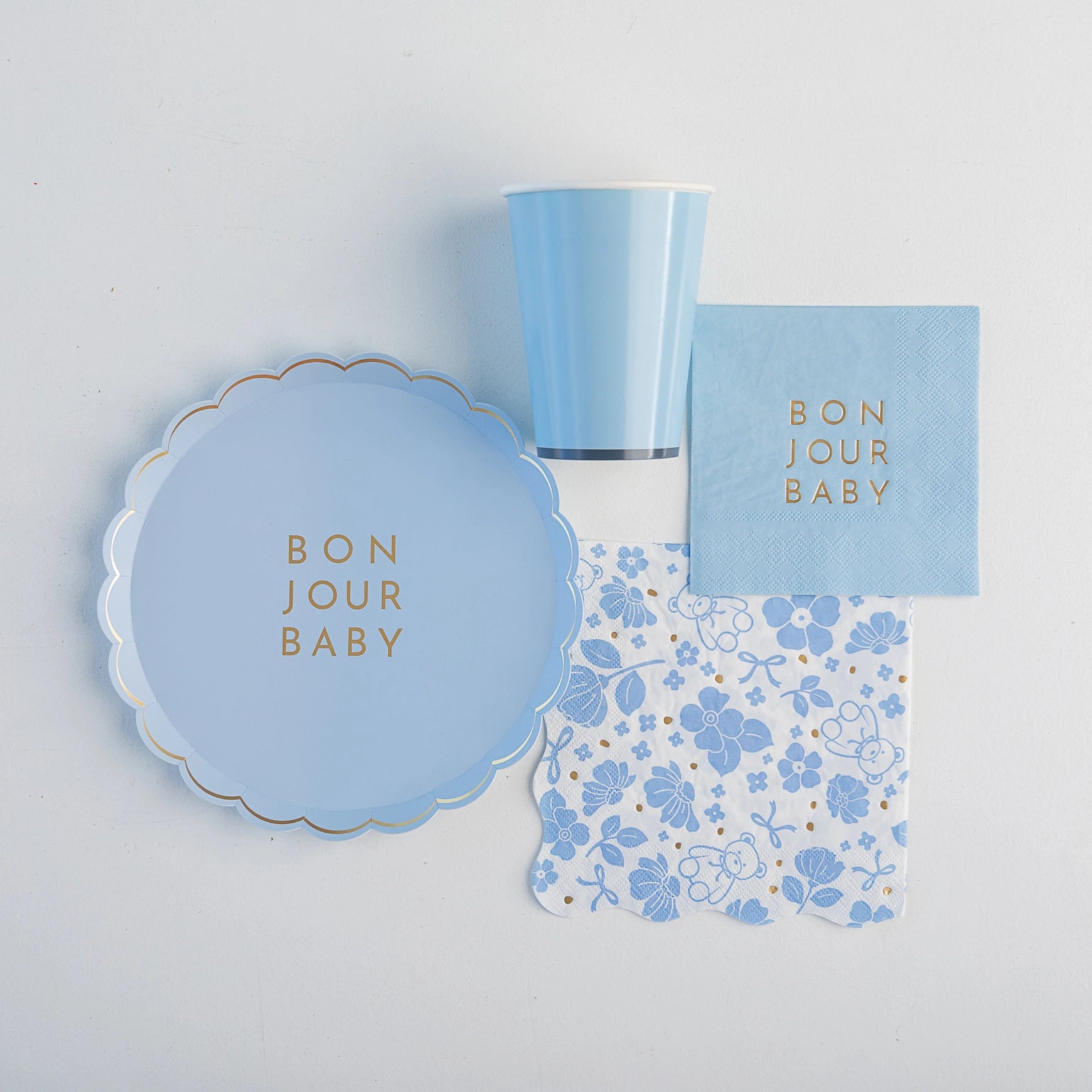 Flat‑lay of Bonjour Baby sky‑blue plate, solid blue cup, gold‑foil cocktail napkin and blue floral lunch napkin on light background.