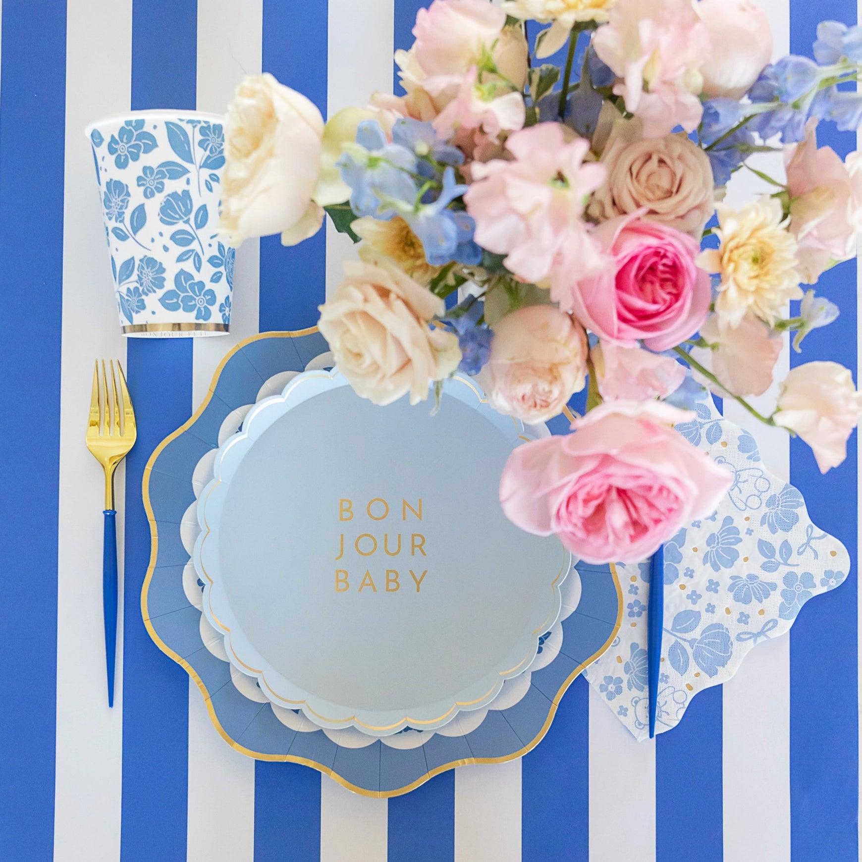 Top‑view tablescape featuring sky‑blue Bonjour Baby plate stack, floral bouquet, blue floral cup and lunch napkin on royal‑blue striped runner.