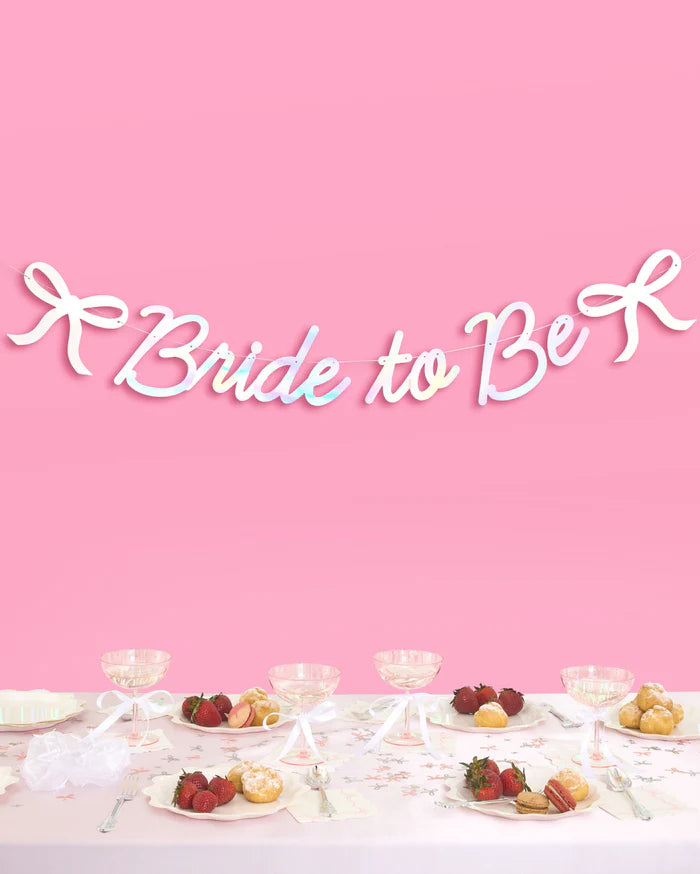 Bride to Be iridescent bow banner hung above bridal shower table with champagne coupes and desserts