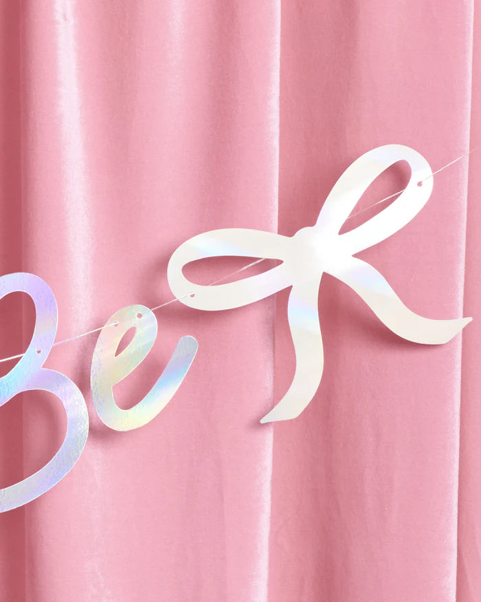 Close-up of iridescent foil bow icon and script lettering on Bride to Be banner against pink velvet