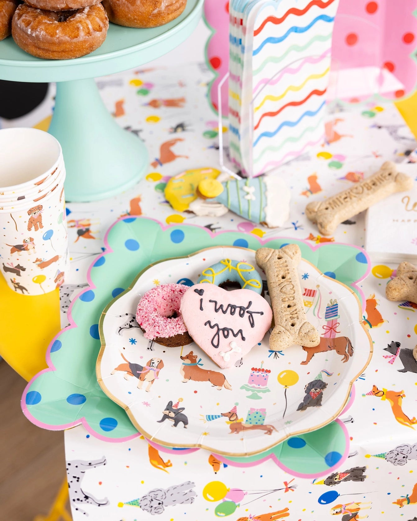 Buffet scene—rainbow guest napkins stacked upright in a clear holder beside donuts, dog treats, and colorful polka-dot plates on a confetti table runner.