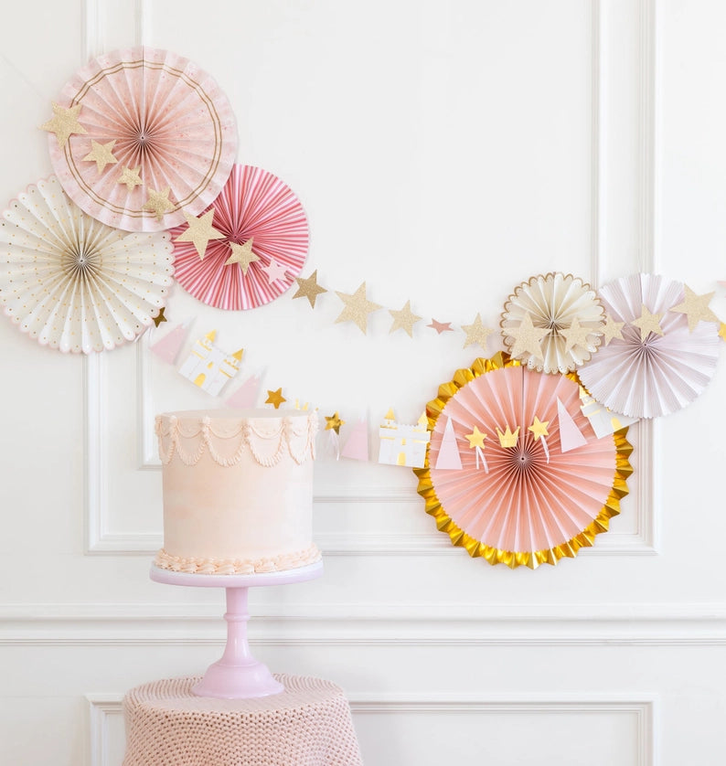 Pink and gold rosette paper fans with glitter-star garland frame a blush cake on a pedestal—perfect princess party decorations backdrop.