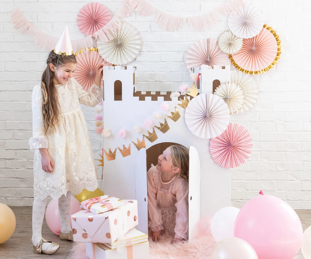 Two children wearing party hats laugh around castle fort draped in gold crowns and pastel pom-pom banner, balloons scattered on floor.