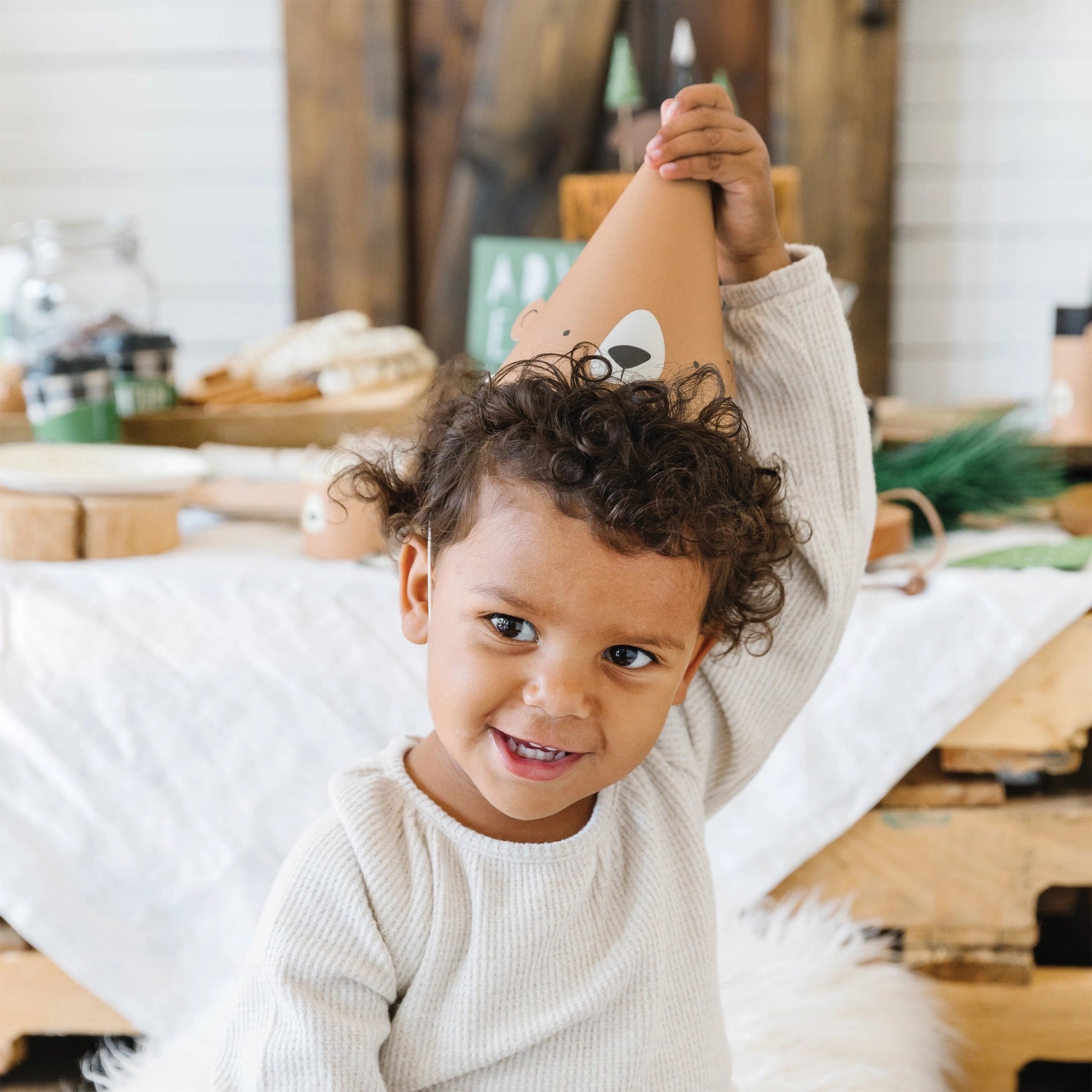 Young child adjusting their bear-themed cone hat while surrounded by rustic camp party decor.