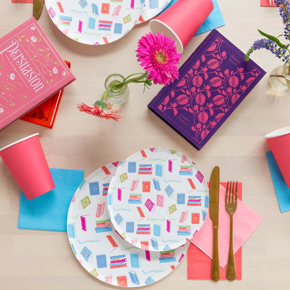 Close view of plates and pink cup beside bright hardcover books and fresh flowers on tabletop.