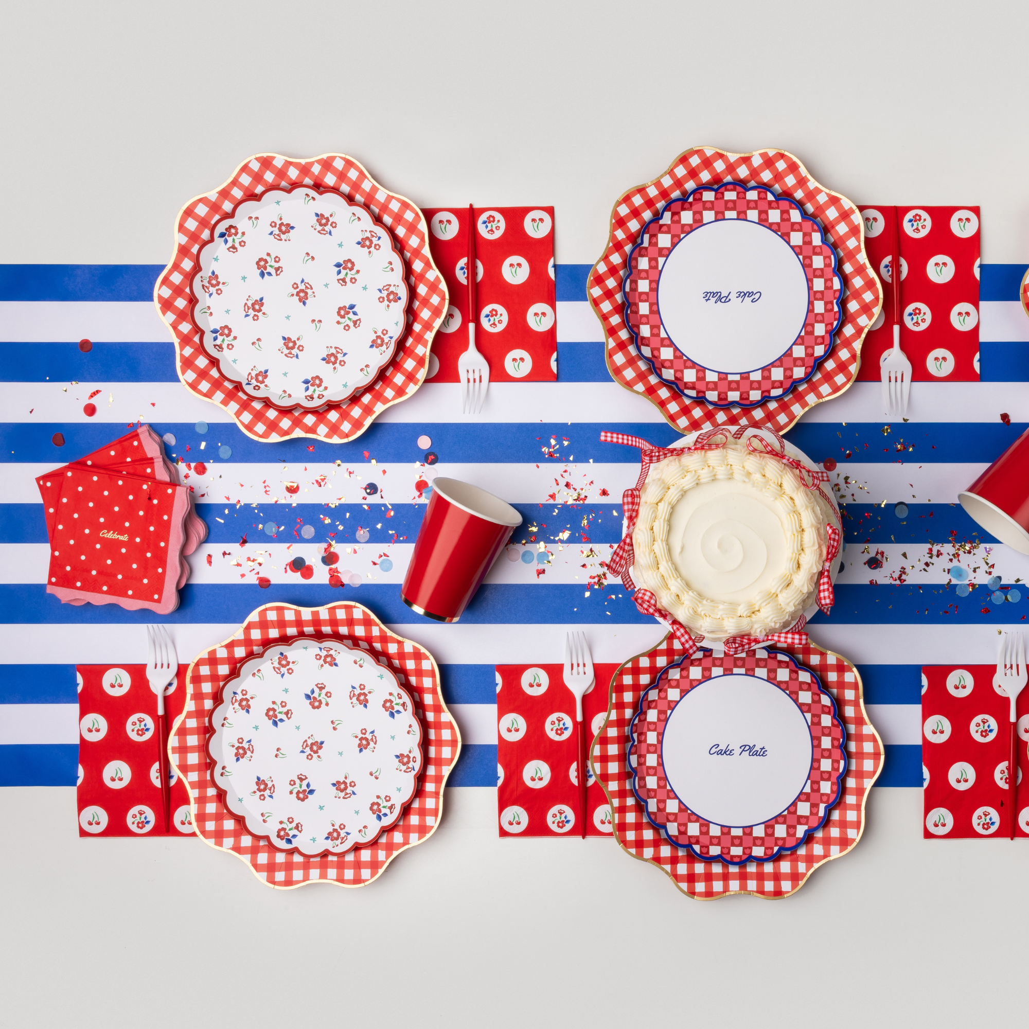 Overhead styled table with red gingham and floral paper plates, cherry print napkins, red polka dot celebrate cocktail napkins, red cups, cake, and blue striped table runner