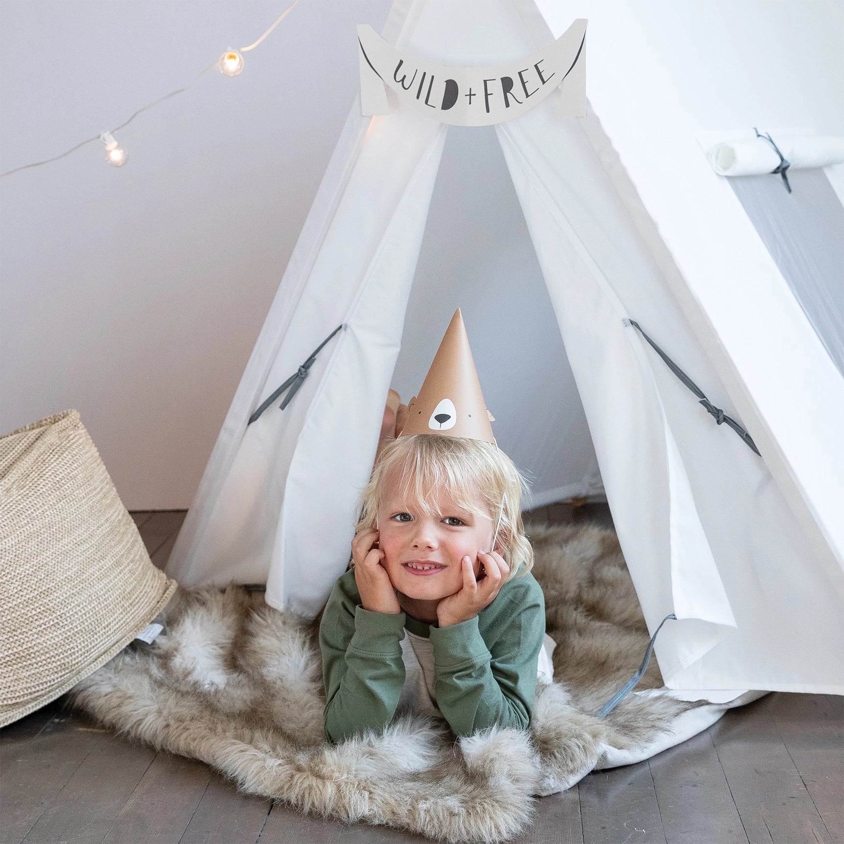 Little camper lying in a teepee wearing the bear hat beneath a “Wild + Free” sign—ideal for a cozy, nature-themed party.