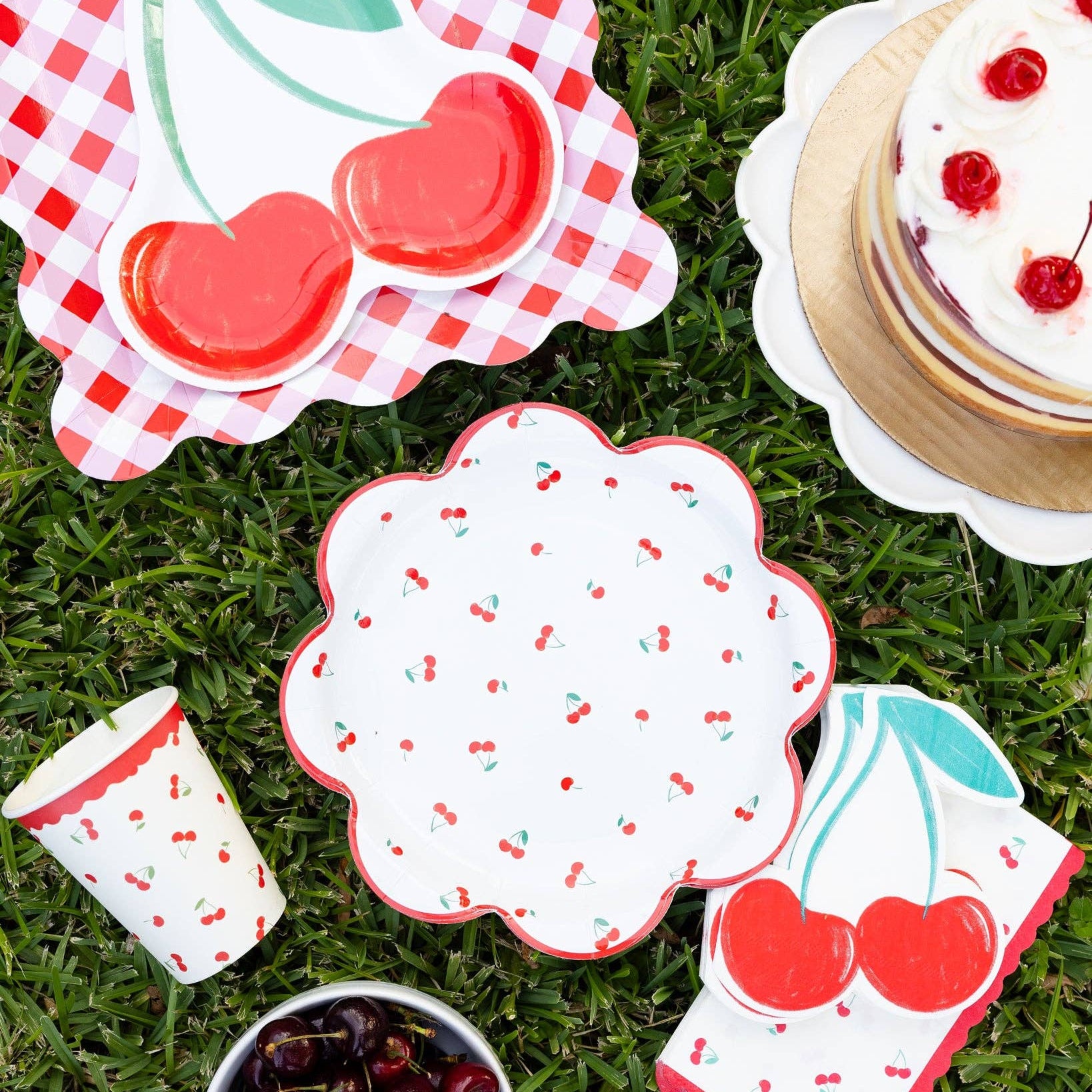Cherry themed party picnic setup with cherry plates, shaped napkins, cake, and cherries on grass.
