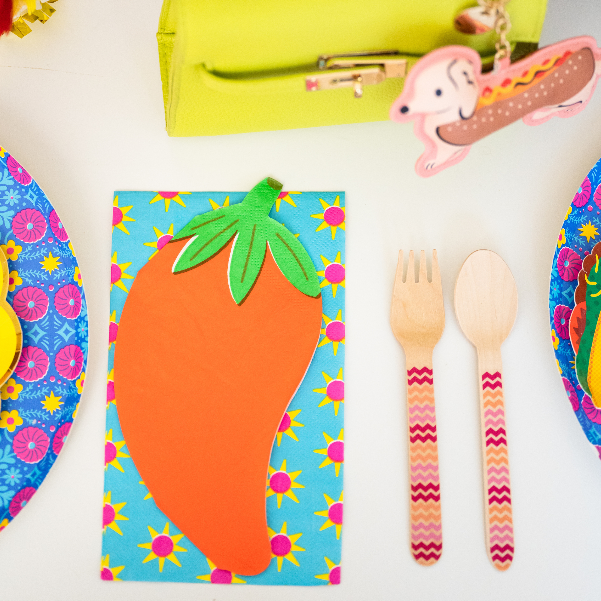 This image shows a chili pepper napkin placed on a colorful taco-themed party table next to wooden utensils and patterned plates.
