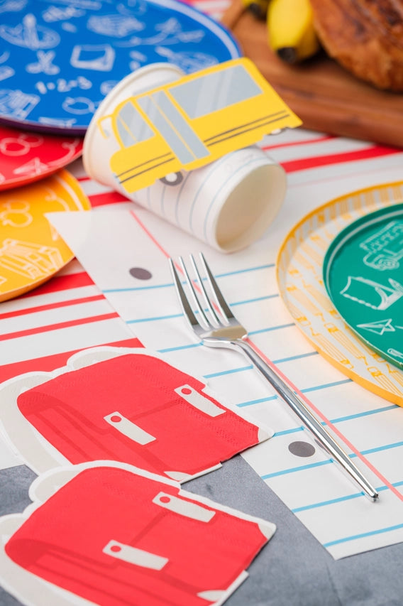 Detail shot of red backpack napkins, fork, and tipped notebook‑paper cup with school‑bus place card on a red‑striped runner and lined placemat.

