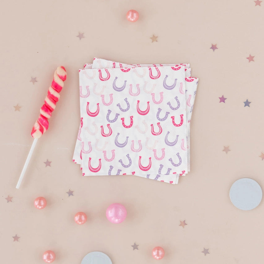 Neatly folded square napkins printed with pink and lavender horseshoes, surrounded by candy pearls, confetti stars, and a pink swirl lollipop.