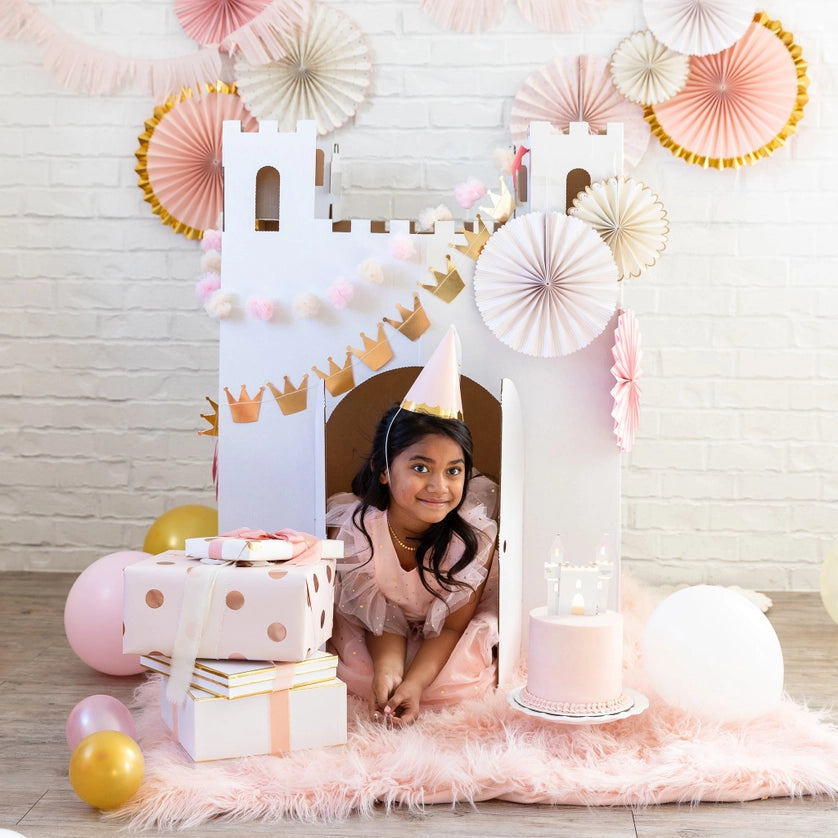 Child in cone hat peeks from a white cardboard castle fort, surrounded by presents, balloons, and pastel rosette fans.
