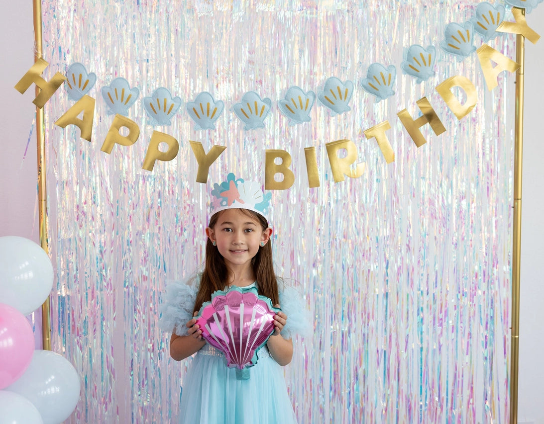 Child in aqua dress holding pink shell balloon beneath seashell and gold “Happy Birthday” banners with iridescent backdrop.
