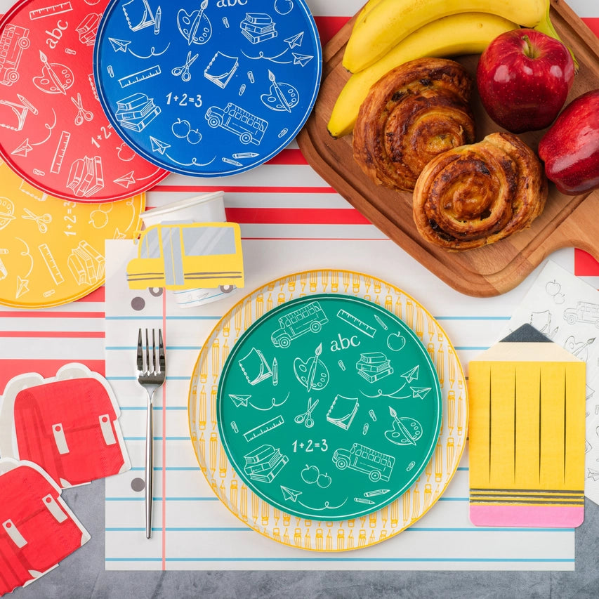 Overhead scene with green doodle plate on yellow pencil‑pattern charger, surrounded by colorful school‑icon plates, pastries, fruit, and notebook placemat.