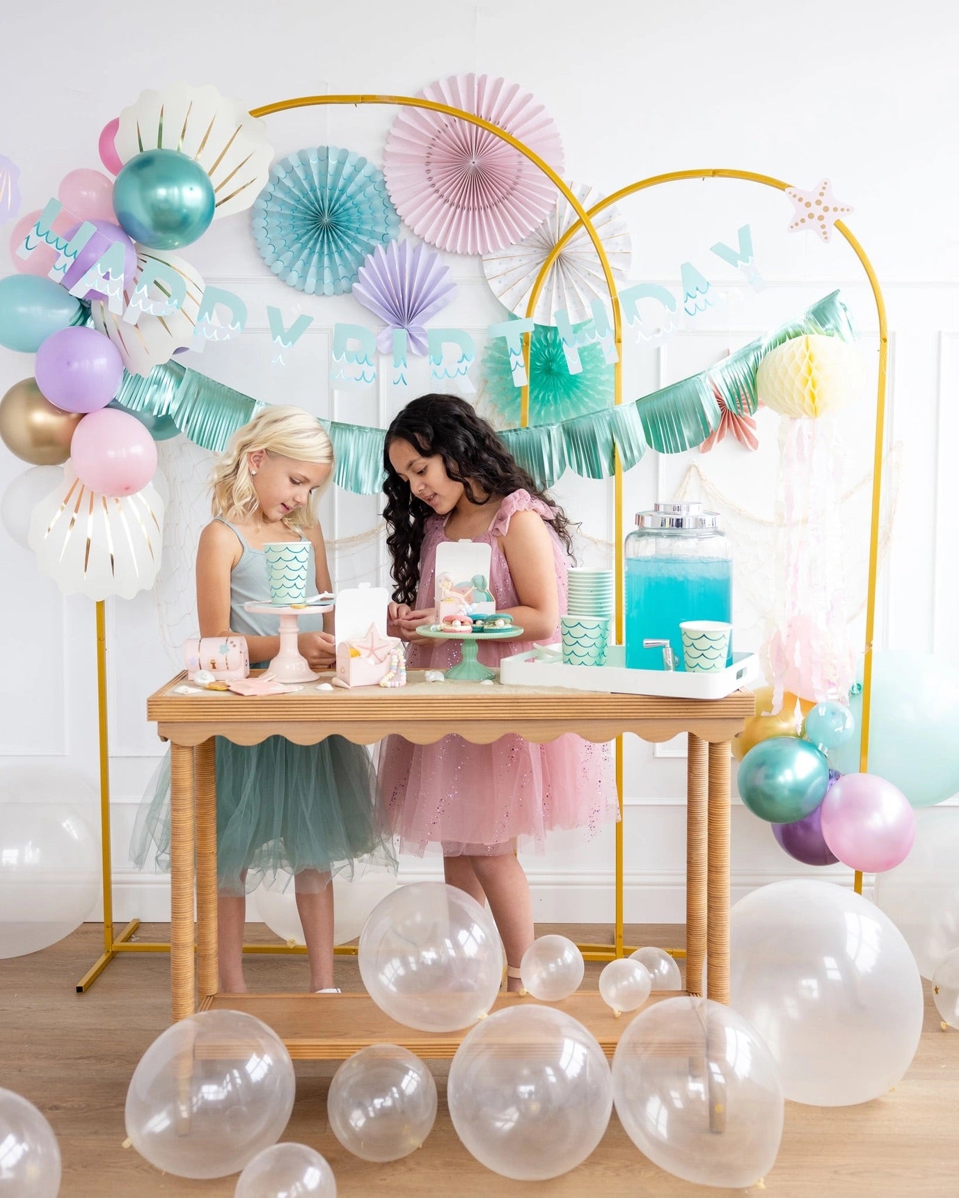 Two little girls dressed in pastel party dresses enjoying treats at a beautifully decorated mermaid-themed party table.
