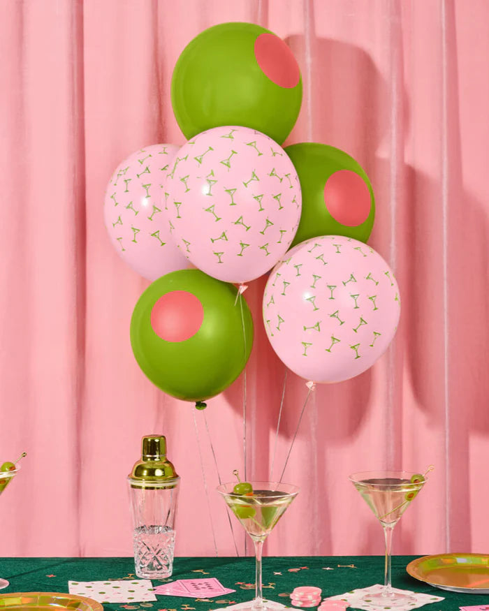 Pink martini and green olive latex balloon bouquet floating above a styled bar table with cocktail shaker, martini glasses, and pink curtain backdrop