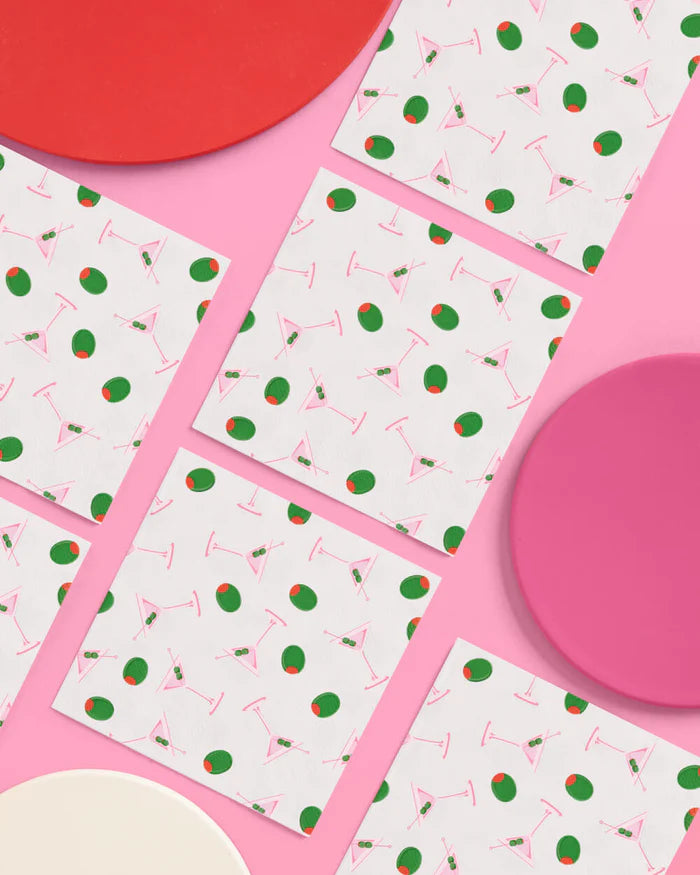 Martini print paper cocktail napkins arranged in a grid with pink and red plates on a pink background