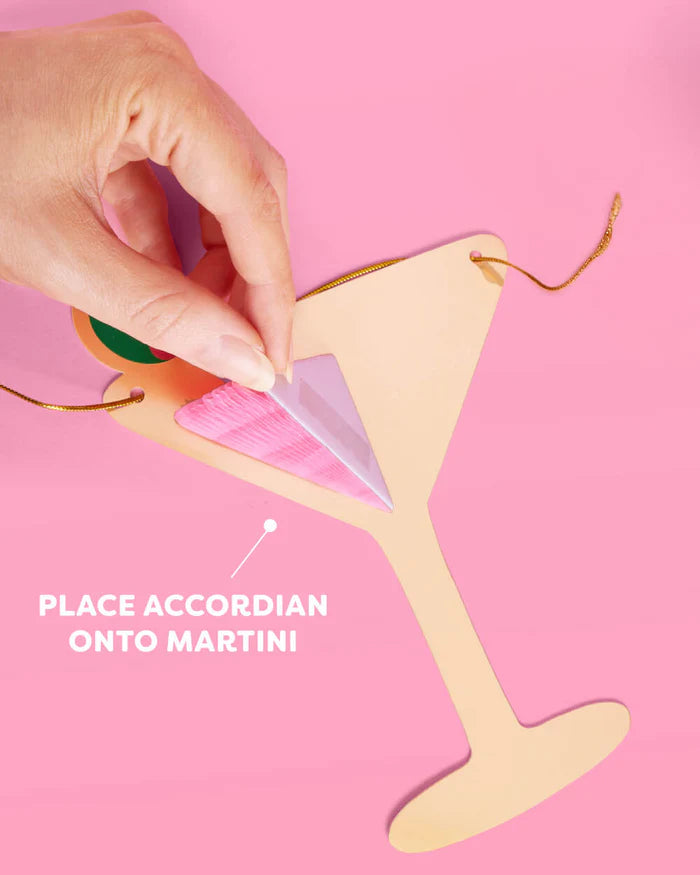 Hand attaching a pink accordion honeycomb insert onto a gold foil die-cut martini glass, showing the assembly step for the Dirty Thirty birthday banner