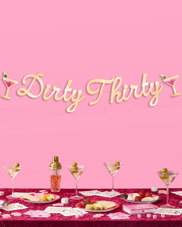 Dirty Thirty gold foil script banner with pink martini glass accents hanging above a styled party table with martini glasses, a cocktail shaker, and charcuterie on a burgundy velvet tablecloth