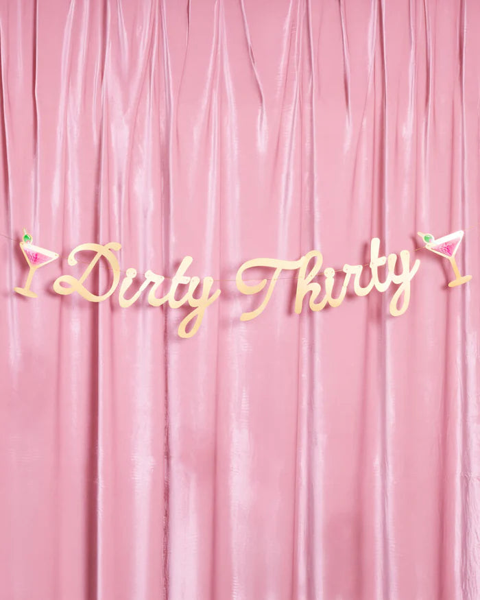 Full Dirty Thirty gold foil script banner with martini glass accents displayed against a pink velvet draped backdrop