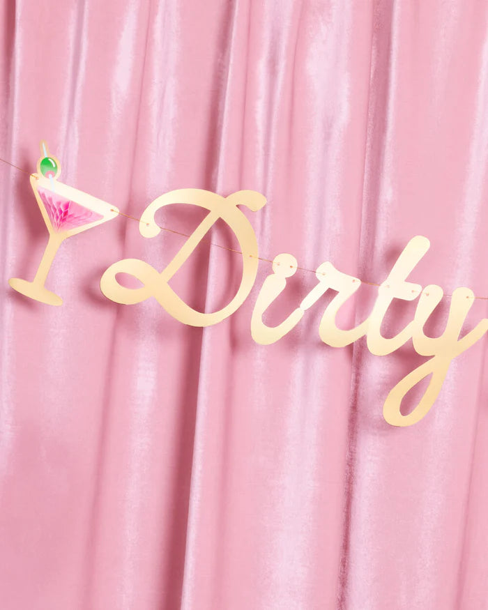 Partial view of gold foil script "Dirty" lettering on the Dirty Thirty birthday banner hanging against a pink velvet drape