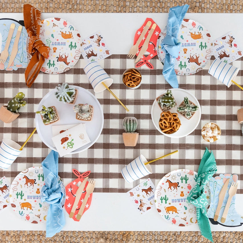 Overhead tablescape: Top‑down view of a Western‑themed kids’ table with cowboy‑illustrated plates, bandana cutlery pockets, mini cacti, and gingham runner.
