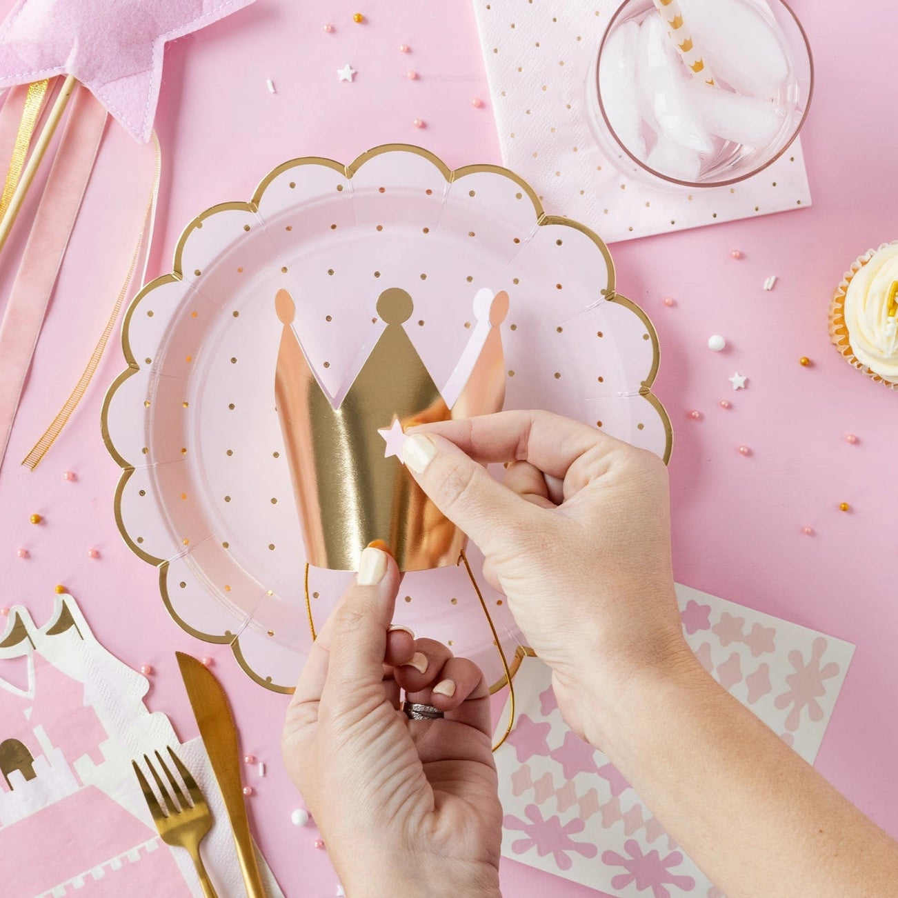 Overhead scene: hands placing star sticker on gold crown plate beside wand, castle napkin, and cupcake.