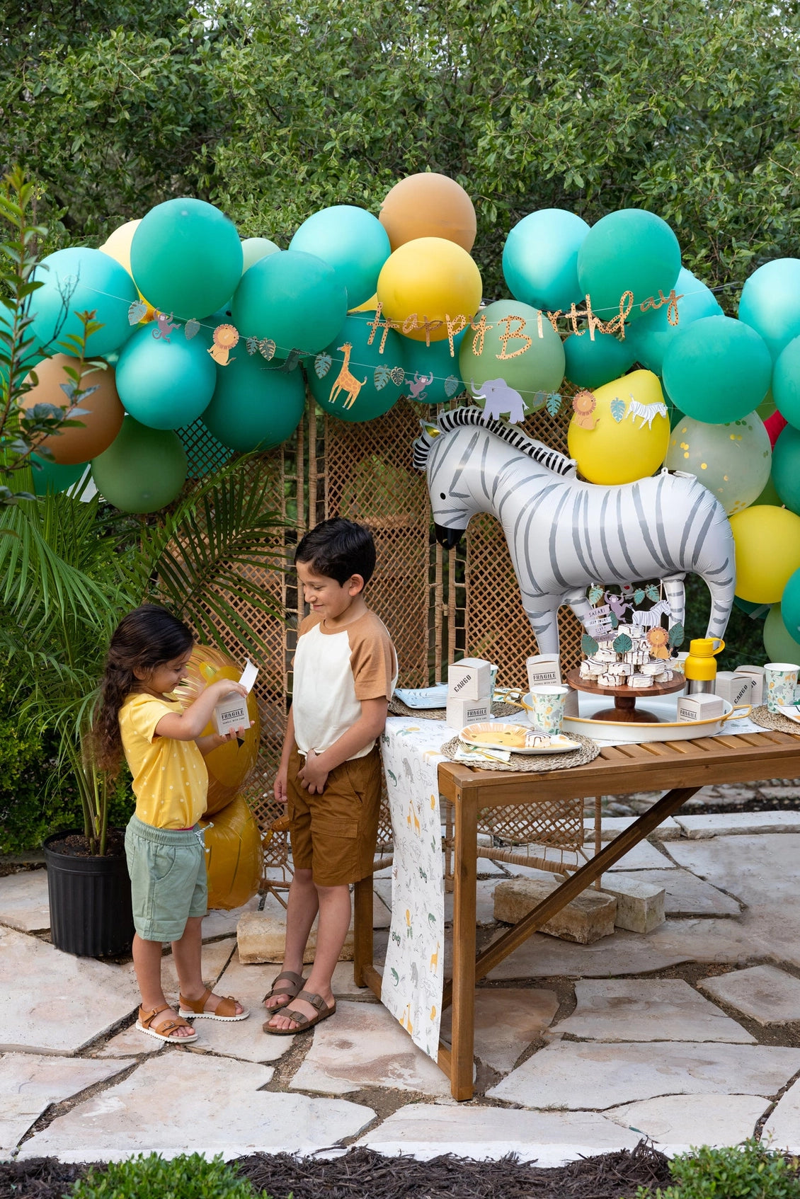 Two kids happily exchanging safari cargo box favors at a festive jungle-themed birthday setup with vibrant balloon arch.