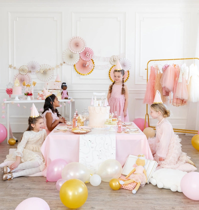 Princess-themed dining scene: girls in party hats smiling at a castle-topped cake, layered paper-fan wall in background.