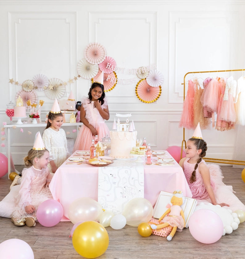 Four girls in pastel dresses gather around a low pink table set for a princess birthday, paper fans and balloons all around.