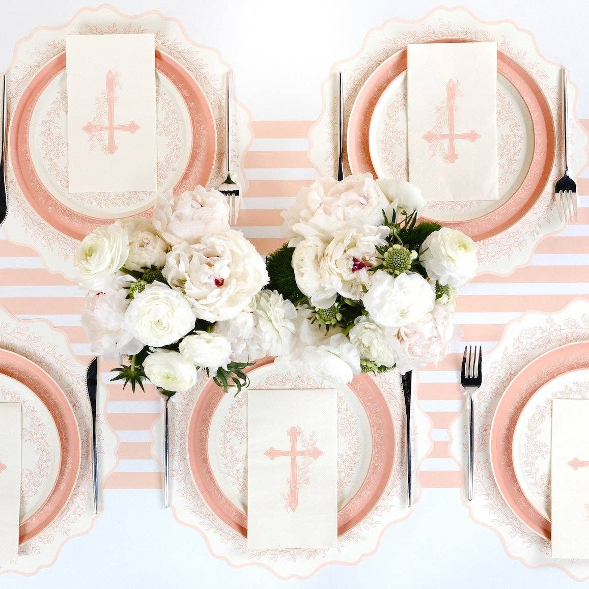 A sunlit English garden baptism table setting featuring botanical cross napkins and pink lattice-edge plates.
