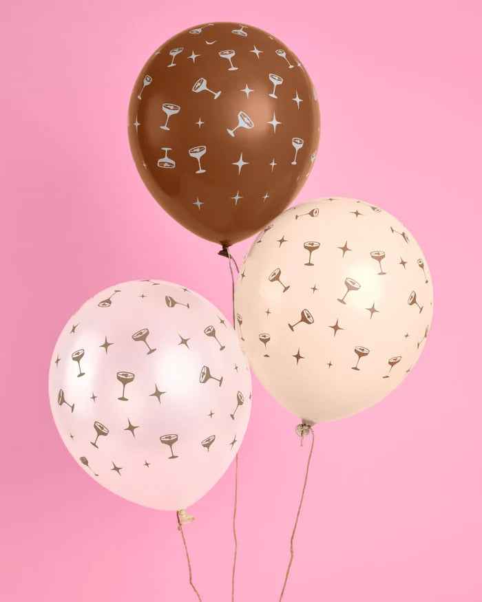 Three matte latex balloons in chocolate brown, blush pink, and cream with an all-over espresso martini coupe and star print, floating against a pink background