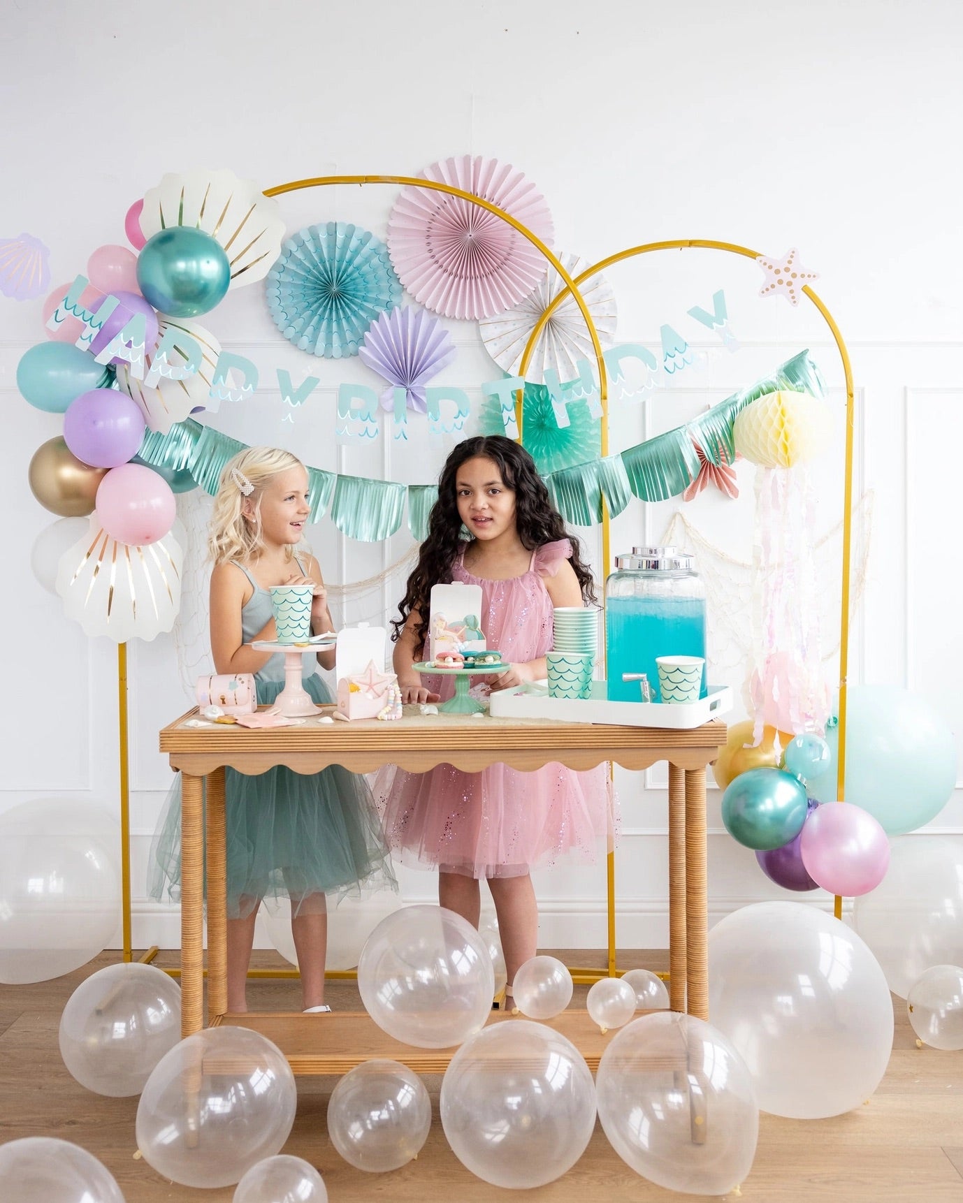 Two young girls chatting and having fun at a mermaid-themed party table, decorated with aqua cups, pastel balloons, and seashell placemats.