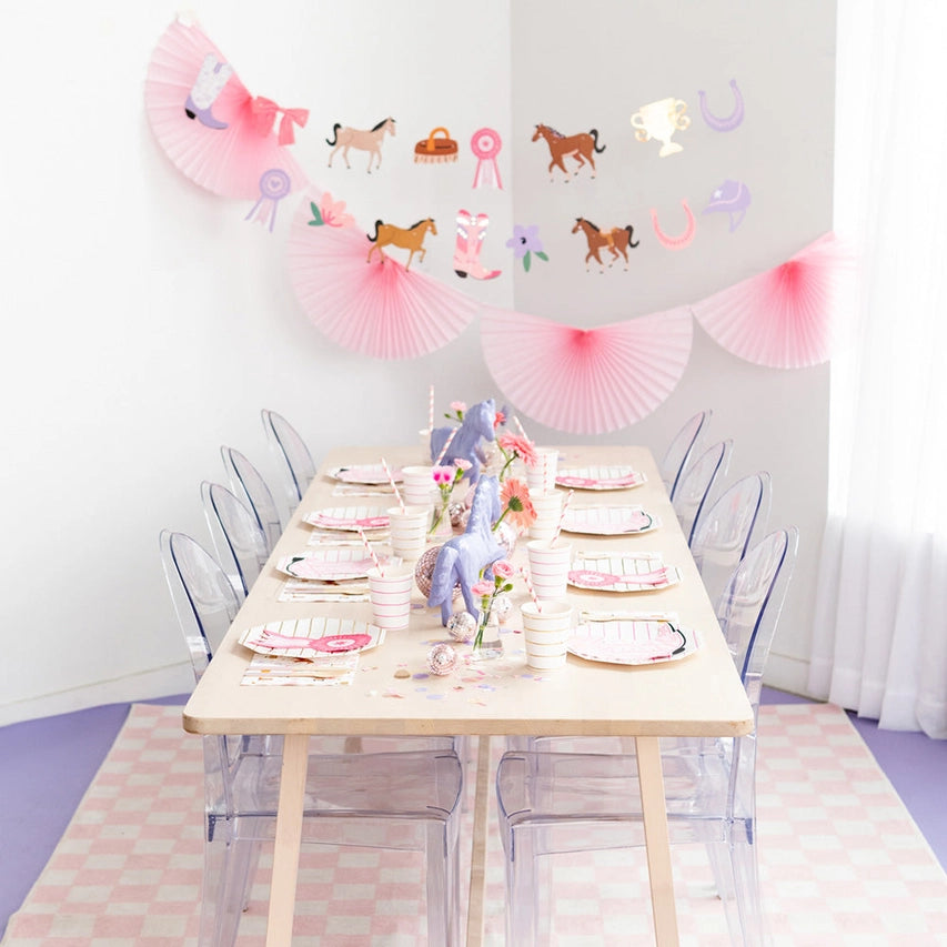 Long wood party table set with Pony Tales plates and cups, purple toy horses as centerpieces, and horse‑themed cut‑out garland draped on white wall.