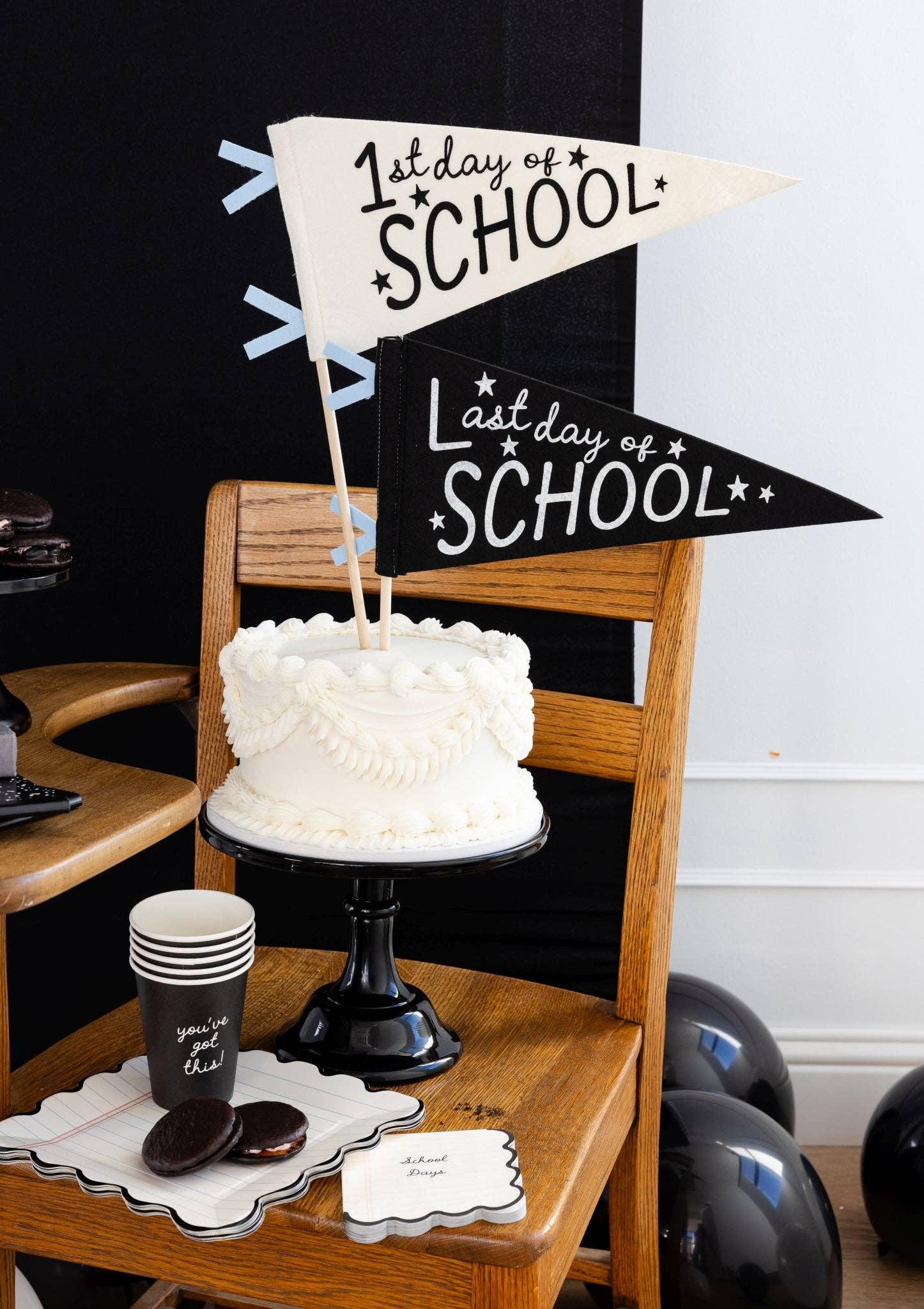 Styled back to school cake table with 1st Day of School and Last Day of School felt pennants as cake toppers on white layer cake, black cake stand, notebook scalloped plates, you've got this paper cup, and chocolate whoopie pies