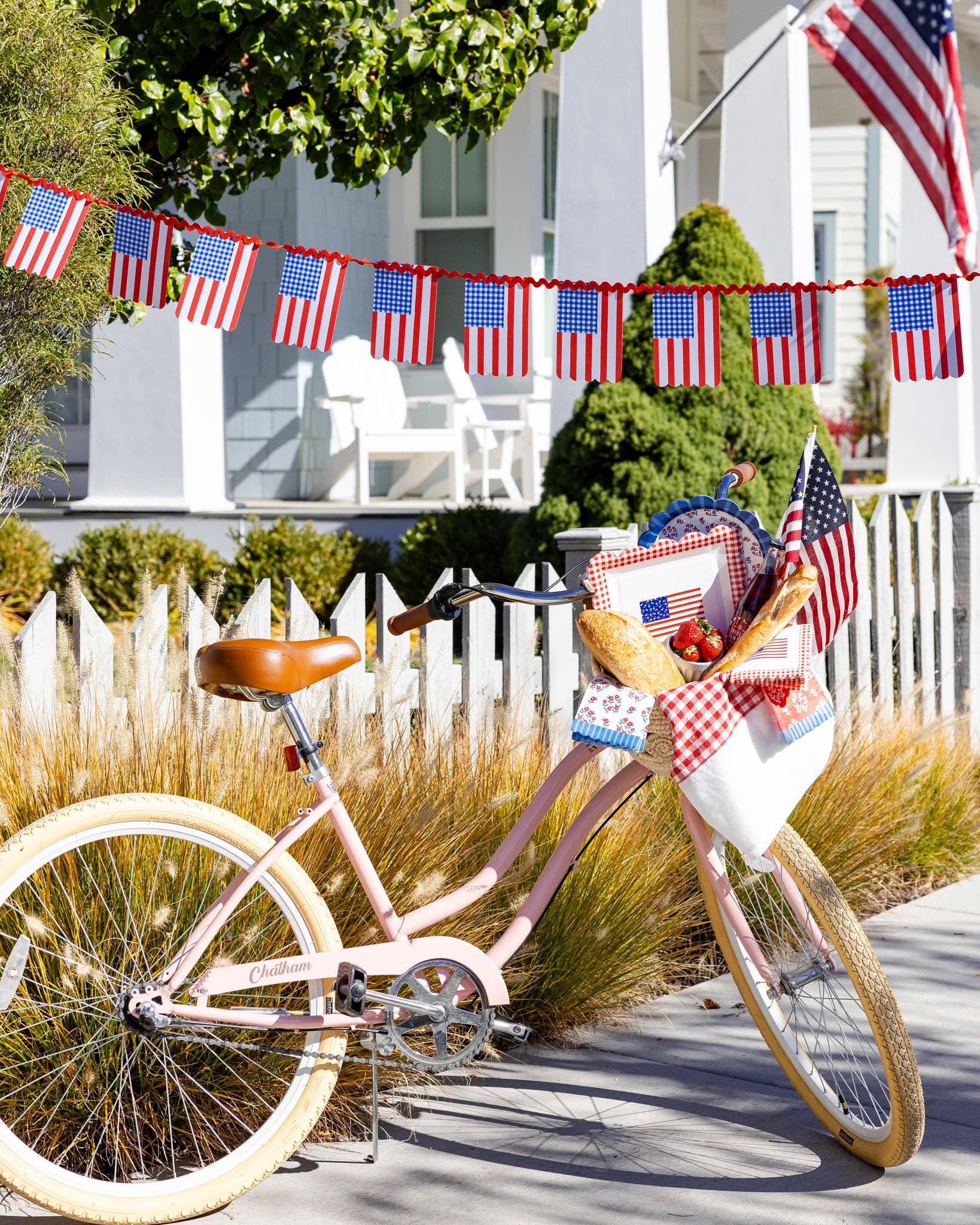 Pink bicycle with basket filled with party supplies and floral napkins in front of white picket fence with American flag bunting
