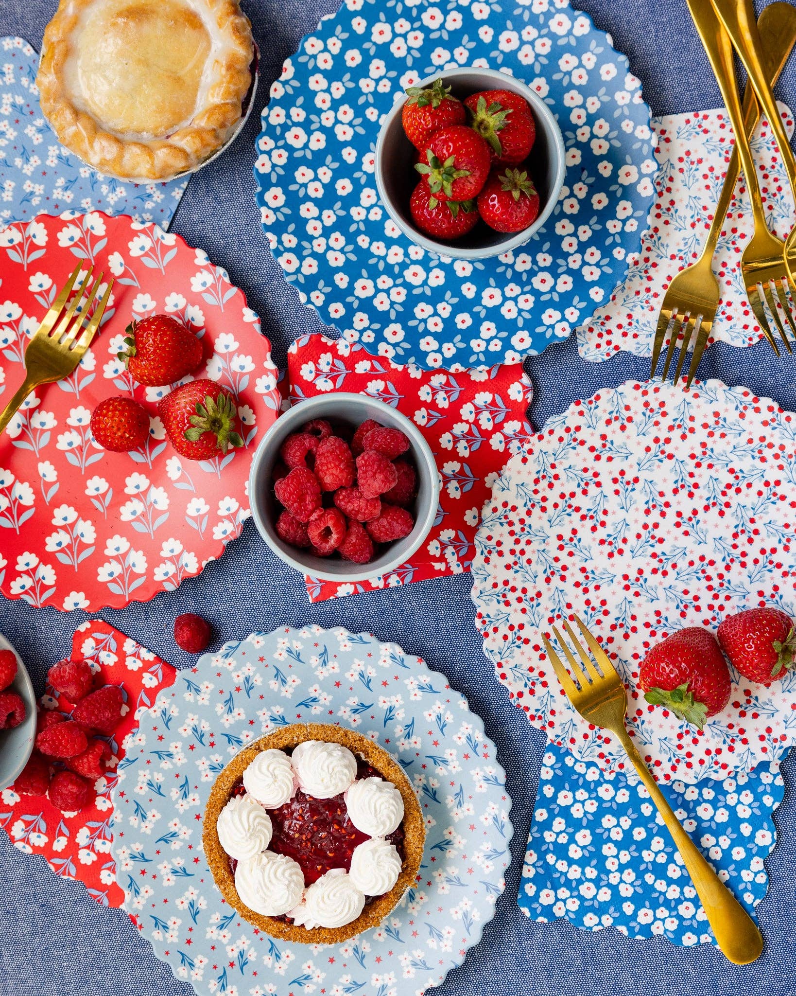 Overhead styled table with all four floral napkin and bamboo plate designs layered with strawberries, raspberries, pie, and gold forks on blue tablecloth