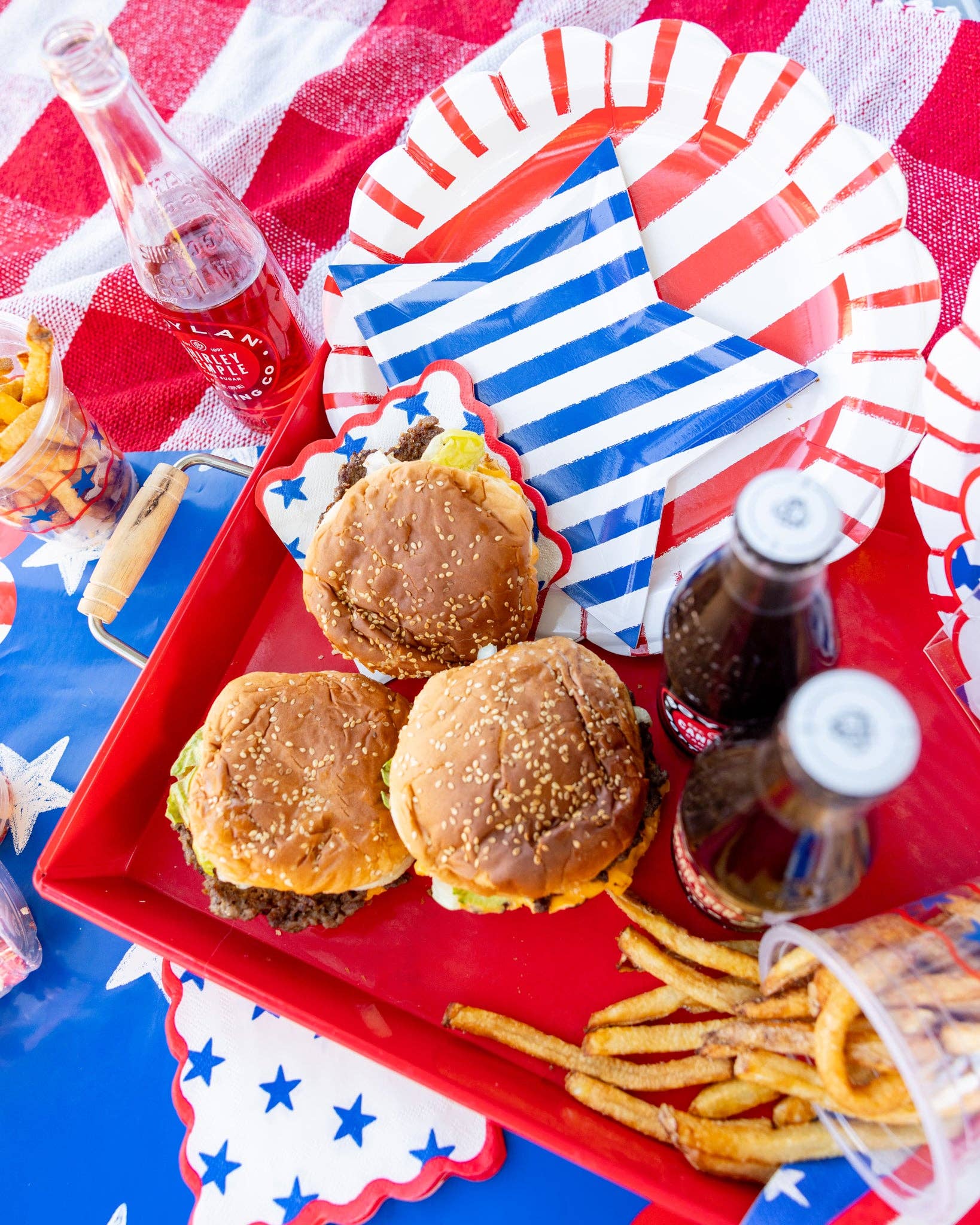 Overhead view of a red tray with cheeseburgers, fries, and bottled sodas alongside blue striped star shaped paper plates and red striped scalloped dinner plates on a patriotic tablescape