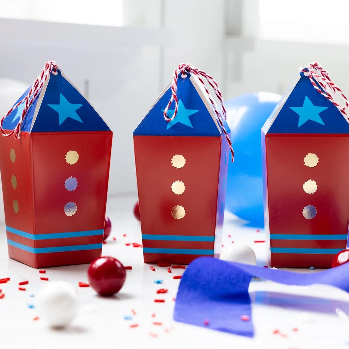 Set of three red, white, and blue rocket-themed treat boxes arranged with party decorations and sweets.